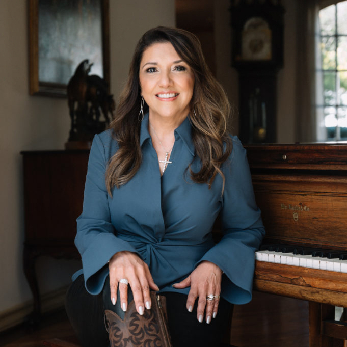 Knight Aerospace CEO Bianca Rhodes seated slightly to the side with a smile as her back is turned to a piano in a home setting. She is wearing a blue shirt, brown leather high top boots, and dark pants. Her wedding ring sparkles almost as bright as her smile.