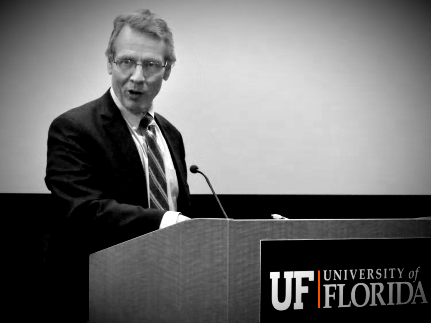 David Parrott at UF speaking and teaching at the podium in black and white with an orange vertical stripe standing out on the front of the wooden podiumj. Dr. Parrott has one hand pointed down and his head cocked to the side above the microphone as he speaks authoritatively.