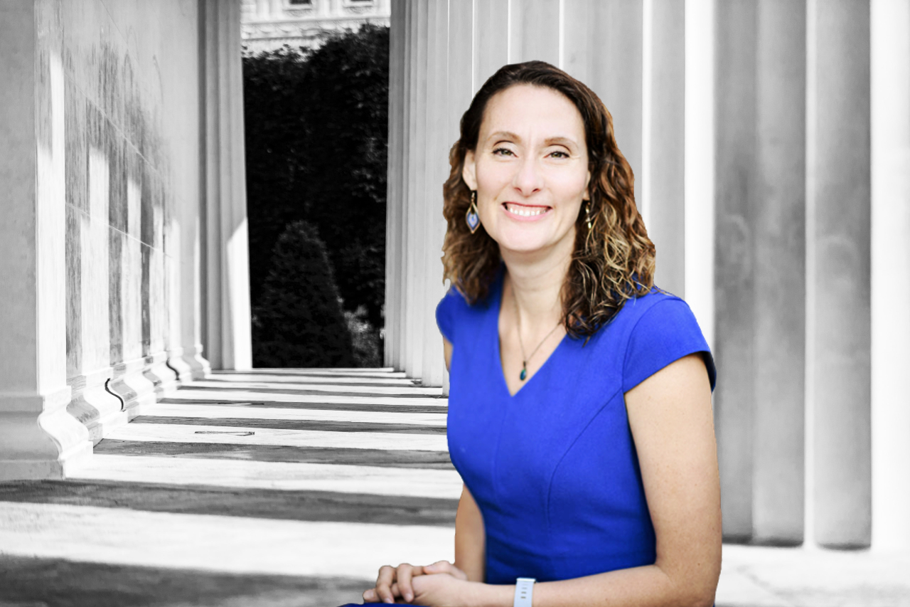 Jessica Dean attorney sitting with hands folded in her lap. She is wearing a blue dress. In the background are Greek columns shown in black and white that look like they belong to a state supreme courthouse.